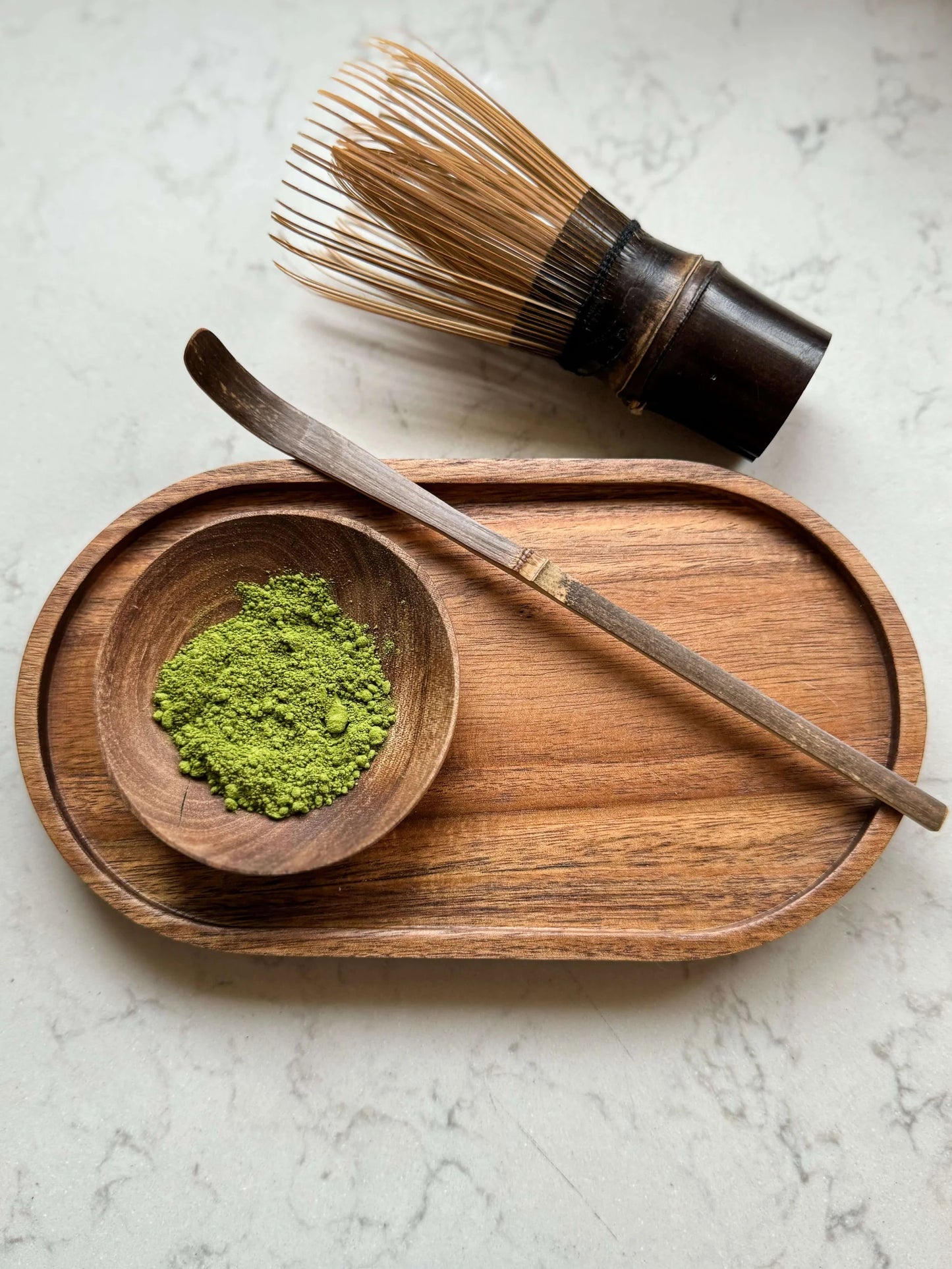 Matcha - Wooden bowl with green powder, wooden whisk, and wooden scoop on a marble surface. Chasen