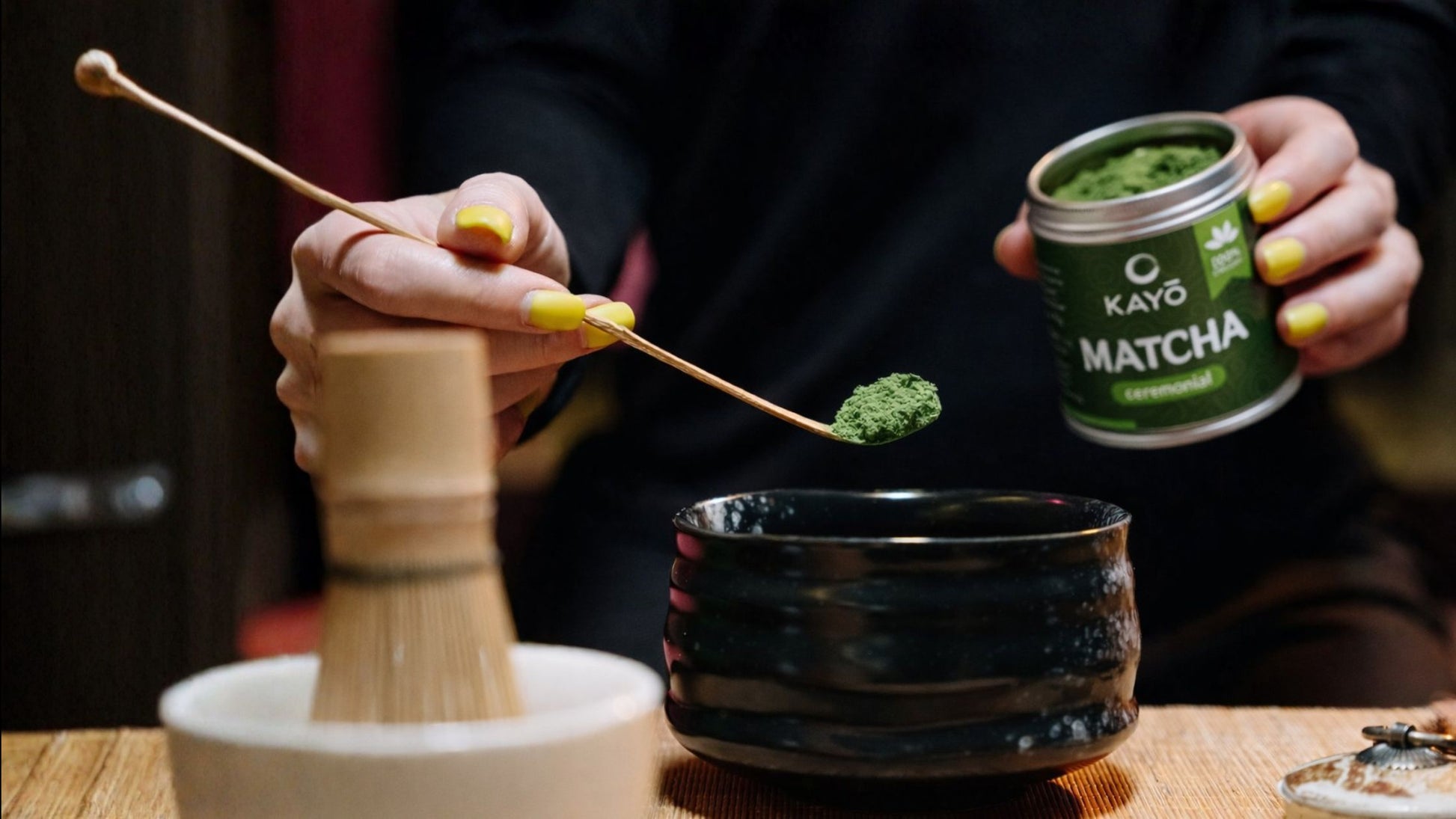 Person preparing matcha with a container of Kayo Matcha on a wooden table.